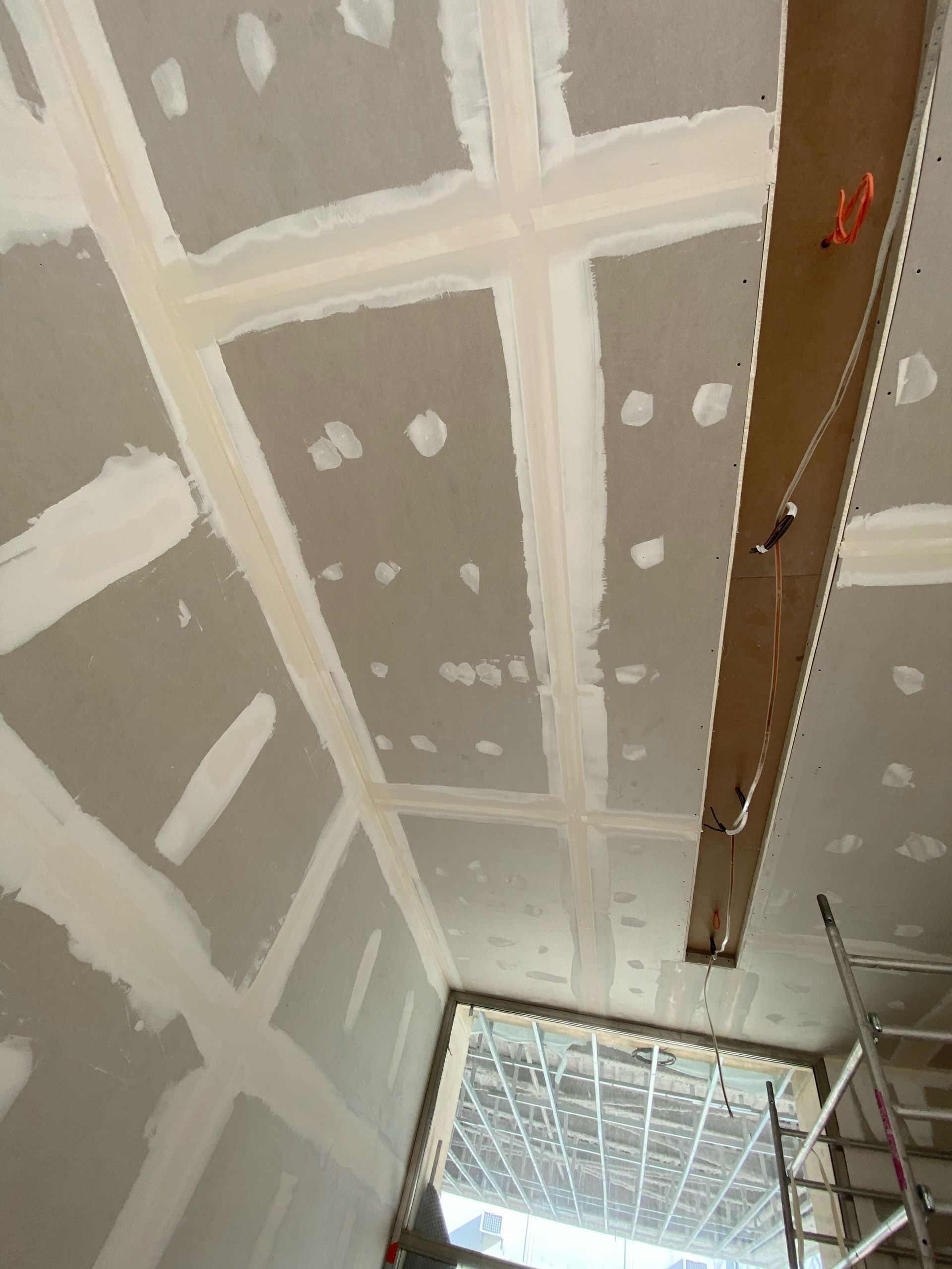 Plafond en plaques de plâtre en cours de construction, avec joints collés, une poutre suspendue et une échelle près d'une fenêtre.
