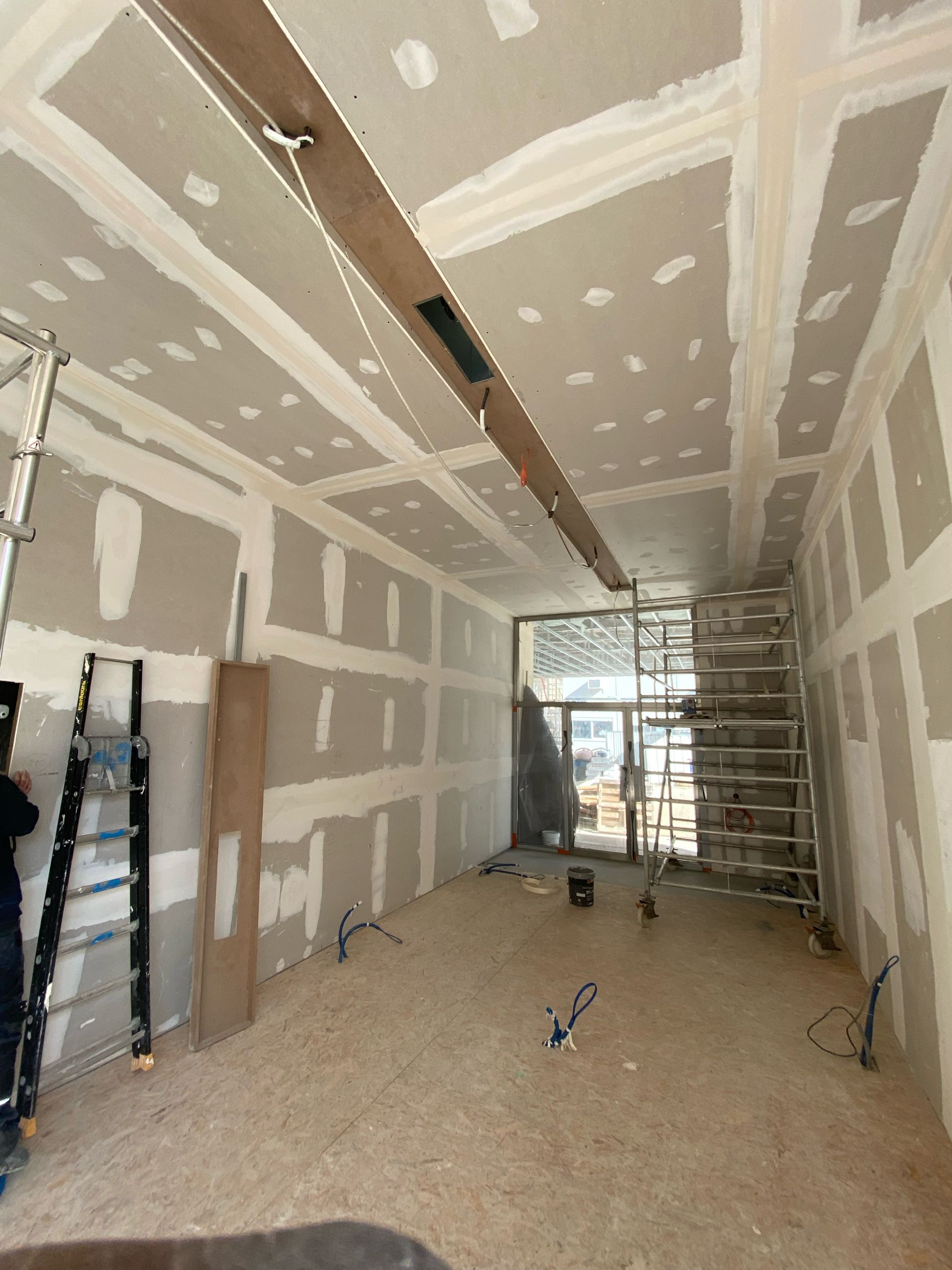 Pièce intérieure recouverte de plaques de plâtre en cours de construction, avec échelles, échafaudages et outils près d'un mur arrière vitré.