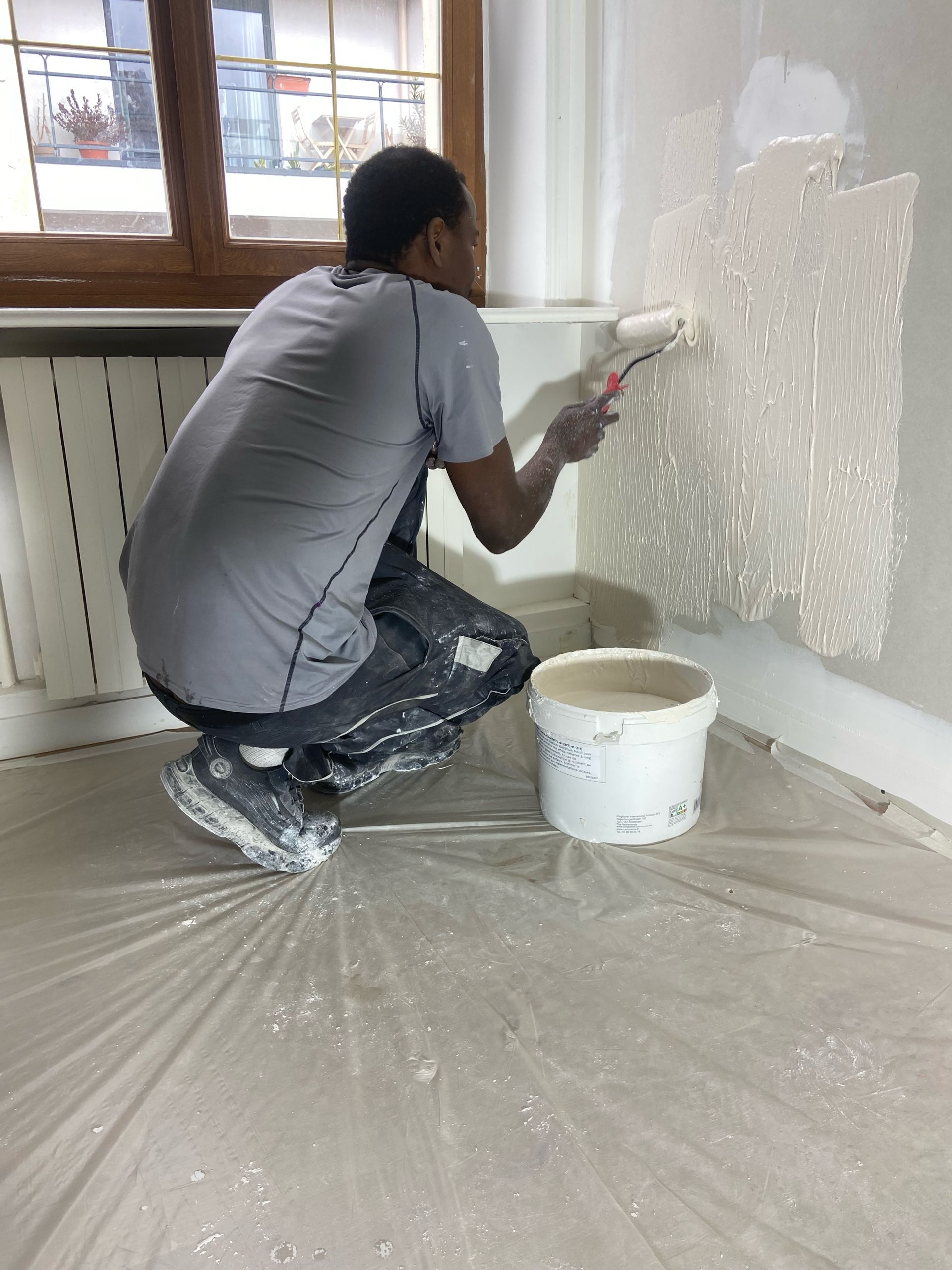 Une personne agenouillée en train de peindre un mur en blanc dans une pièce, avec un seau de peinture posé au sol.