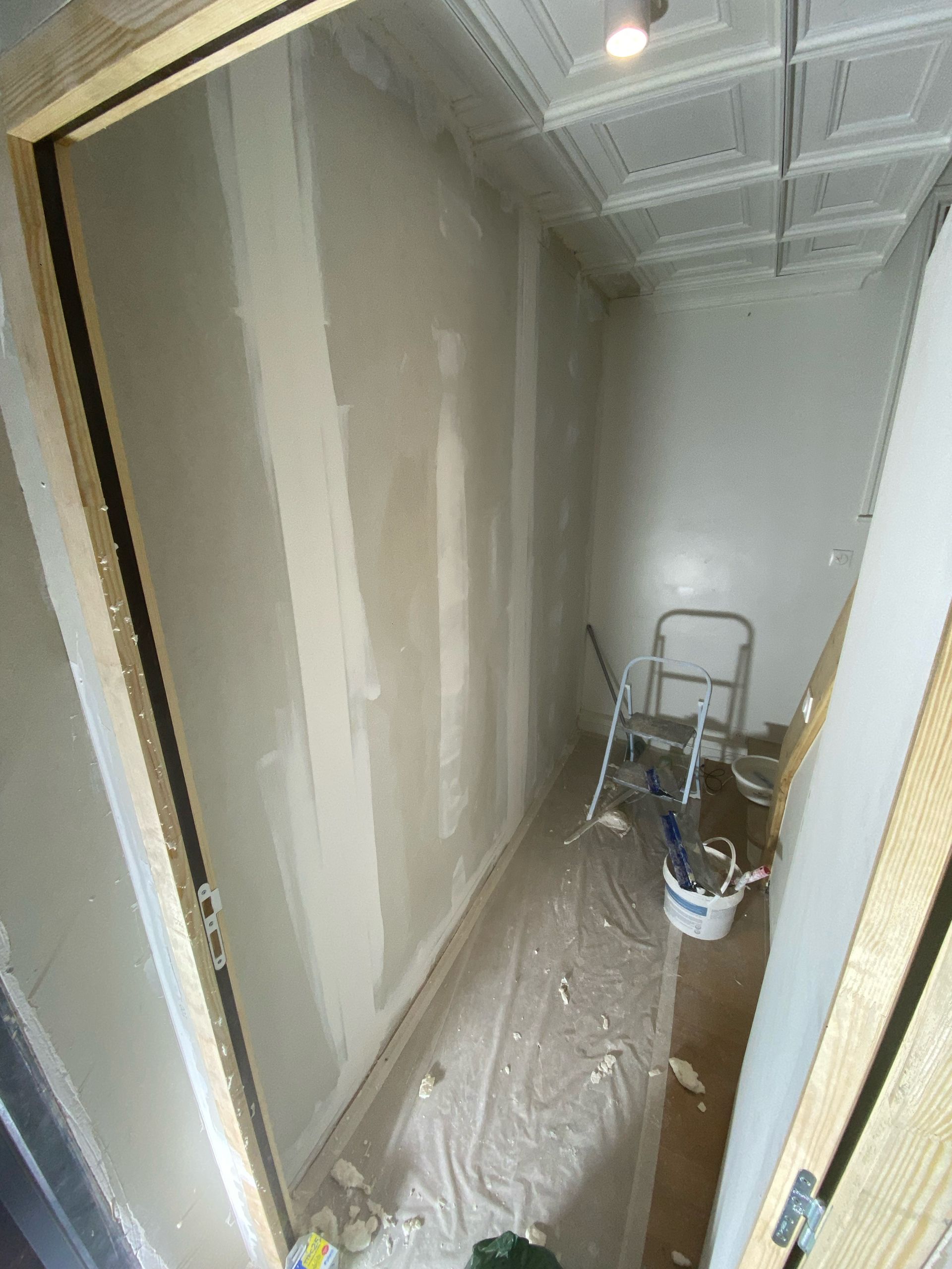 Couloir étroit en cours de rénovation, avec cloisons sèches recouvertes de ruban adhésif, sol recouvert de plastique et une échelle près du fond.
