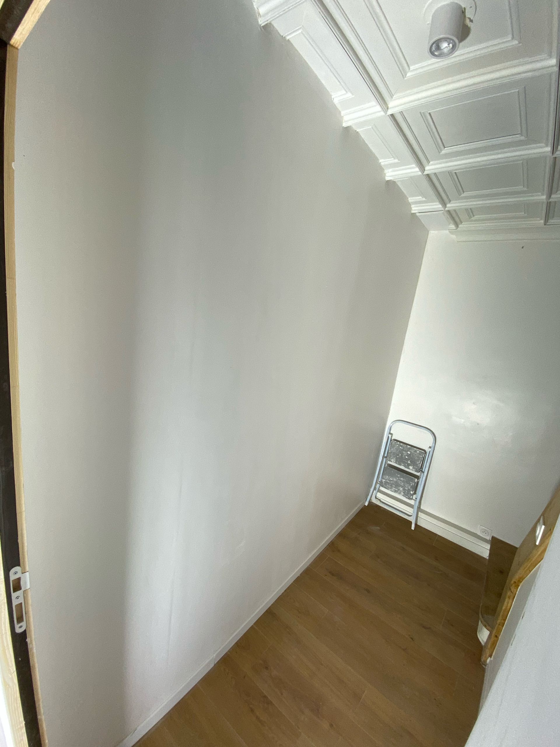 Couloir étroit et blanc avec parquet, plafond orné et une chaise pliante au fond.