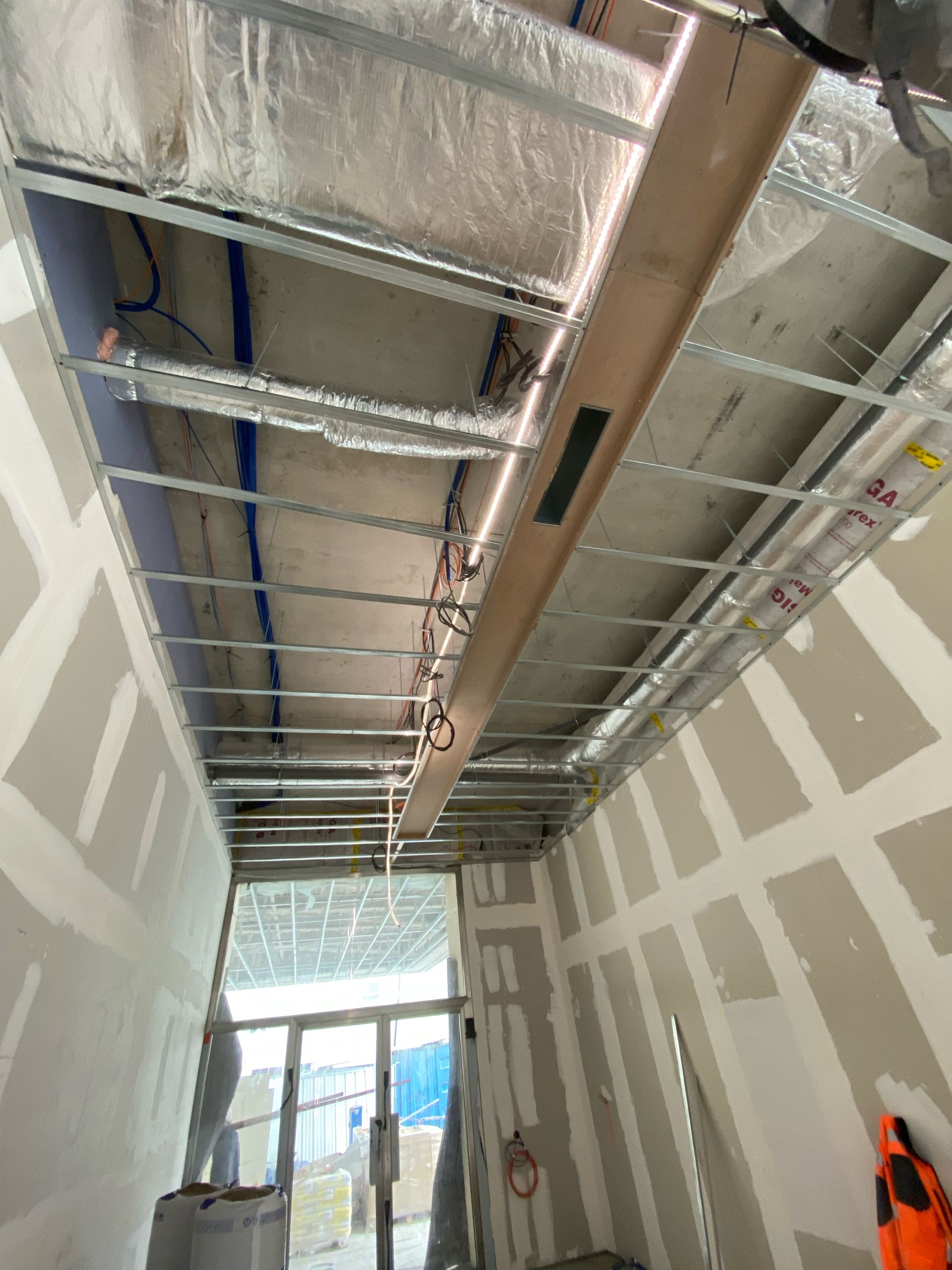 Couloir inachevé avec conduits d'aération apparents au plafond, cloisons sèches et une porte lumineuse au fond.
