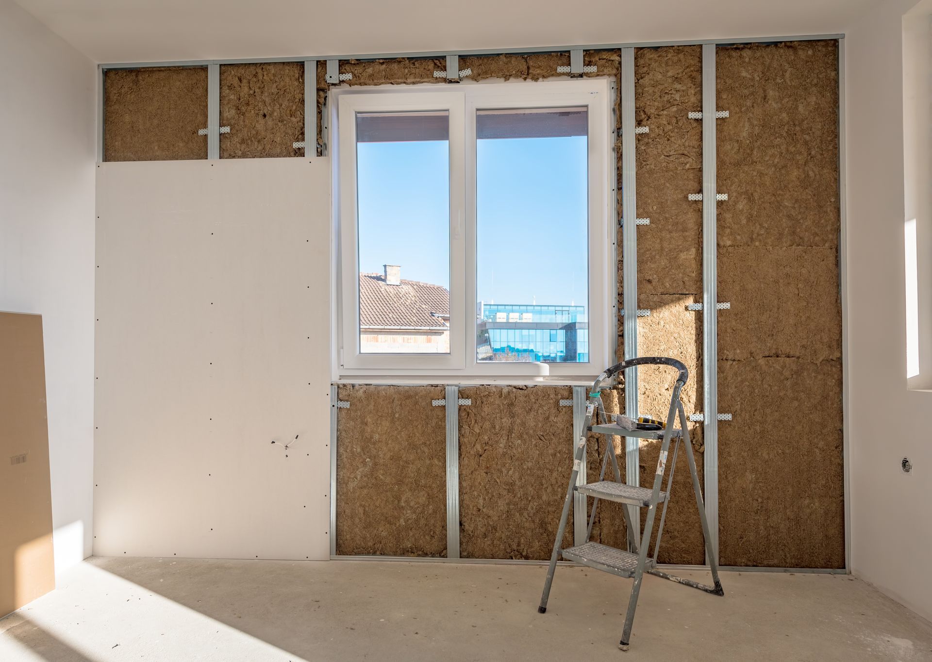Pièce partiellement rénovée avec isolation murale apparente, une fenêtre et un escabeau au sol.