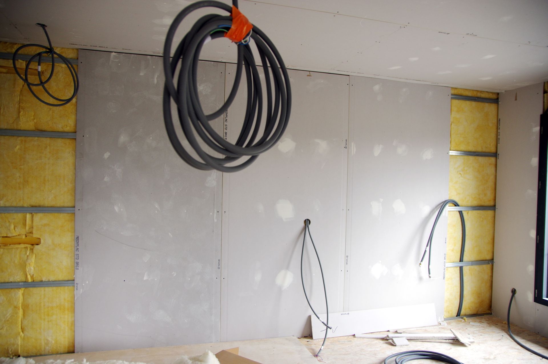 Pièce inachevée avec cloisons sèches, isolation apparente et câbles enroulés pendant du plafond.