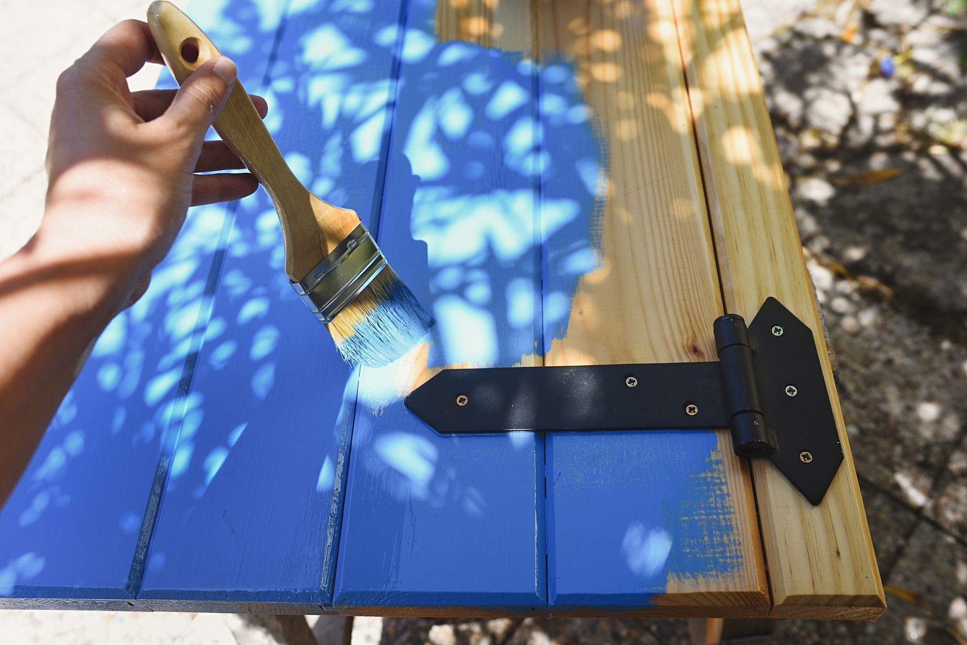 Une main peint un volet en bois bleu avec un pinceau à l'extérieur.