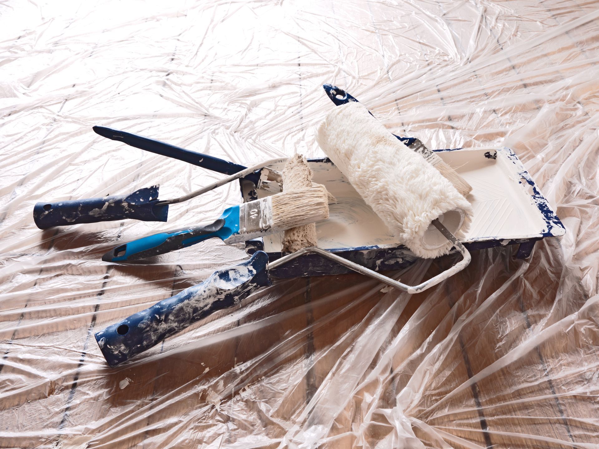 Rouleau à peinture, pinceaux et grattoir sur une bâche en plastique recouverte d'éclaboussures de peinture blanche.