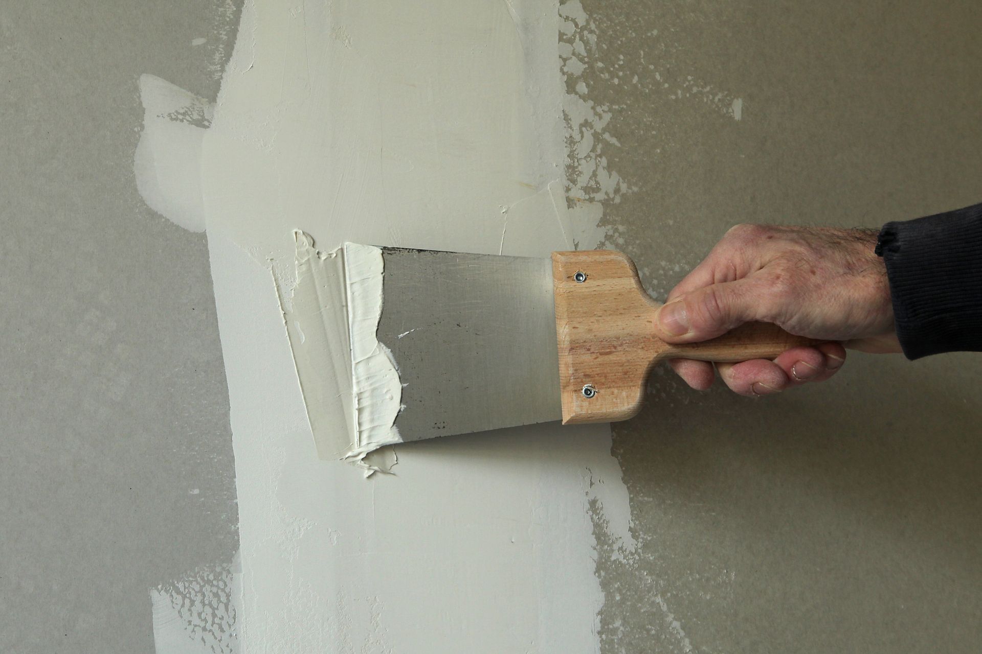 Une main applique du mastic blanc sur un mur gris à l'aide d'un large couteau à mastic.