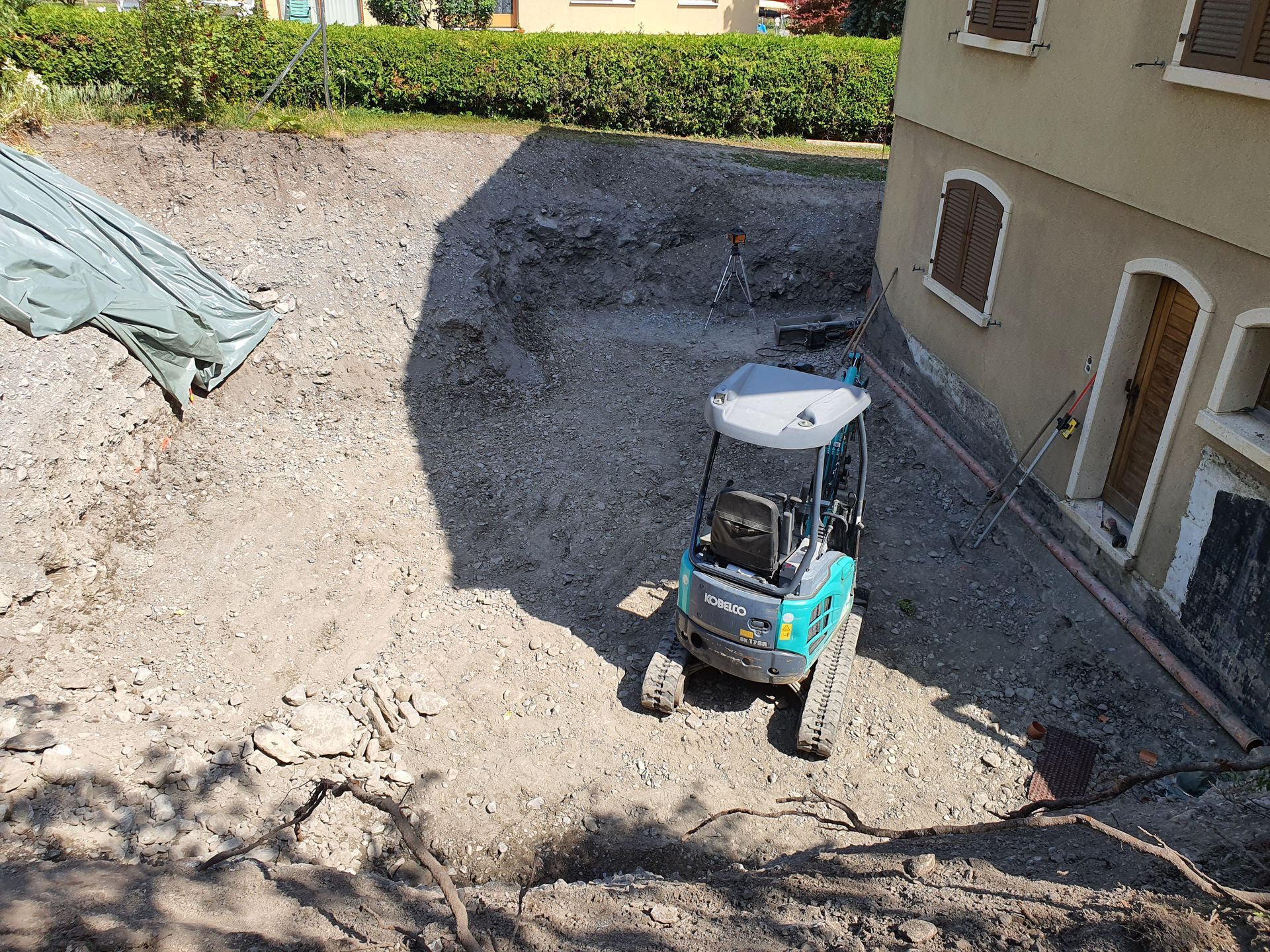 Petite pelleteuse bleue dans une cour en terre battue à côté d'une maison, avec des monticules de gravier et de terre