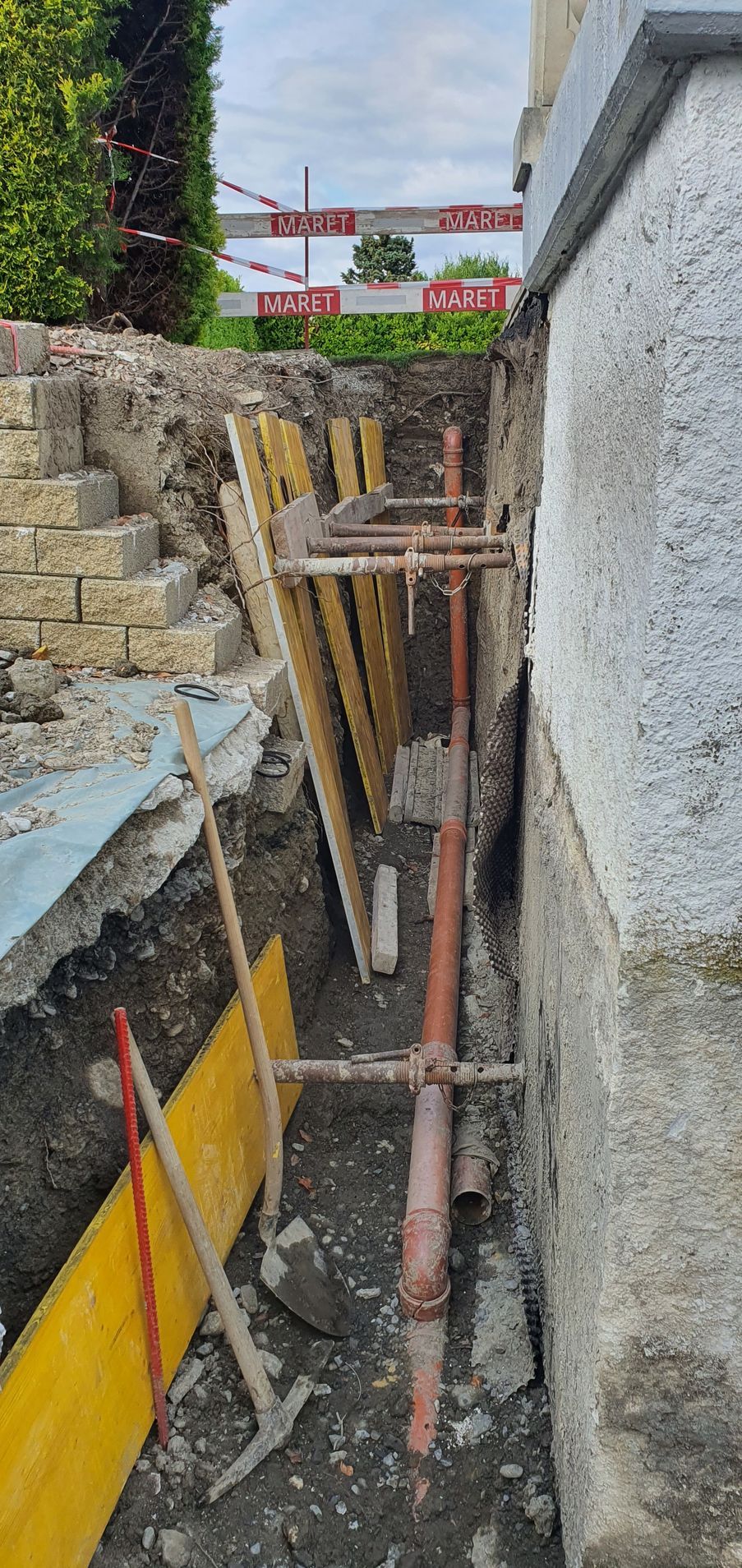 Tranchée de construction étroite avec supports en bois, tuyaux et murs de soutènement en pierre à côté d'un bâtiment.