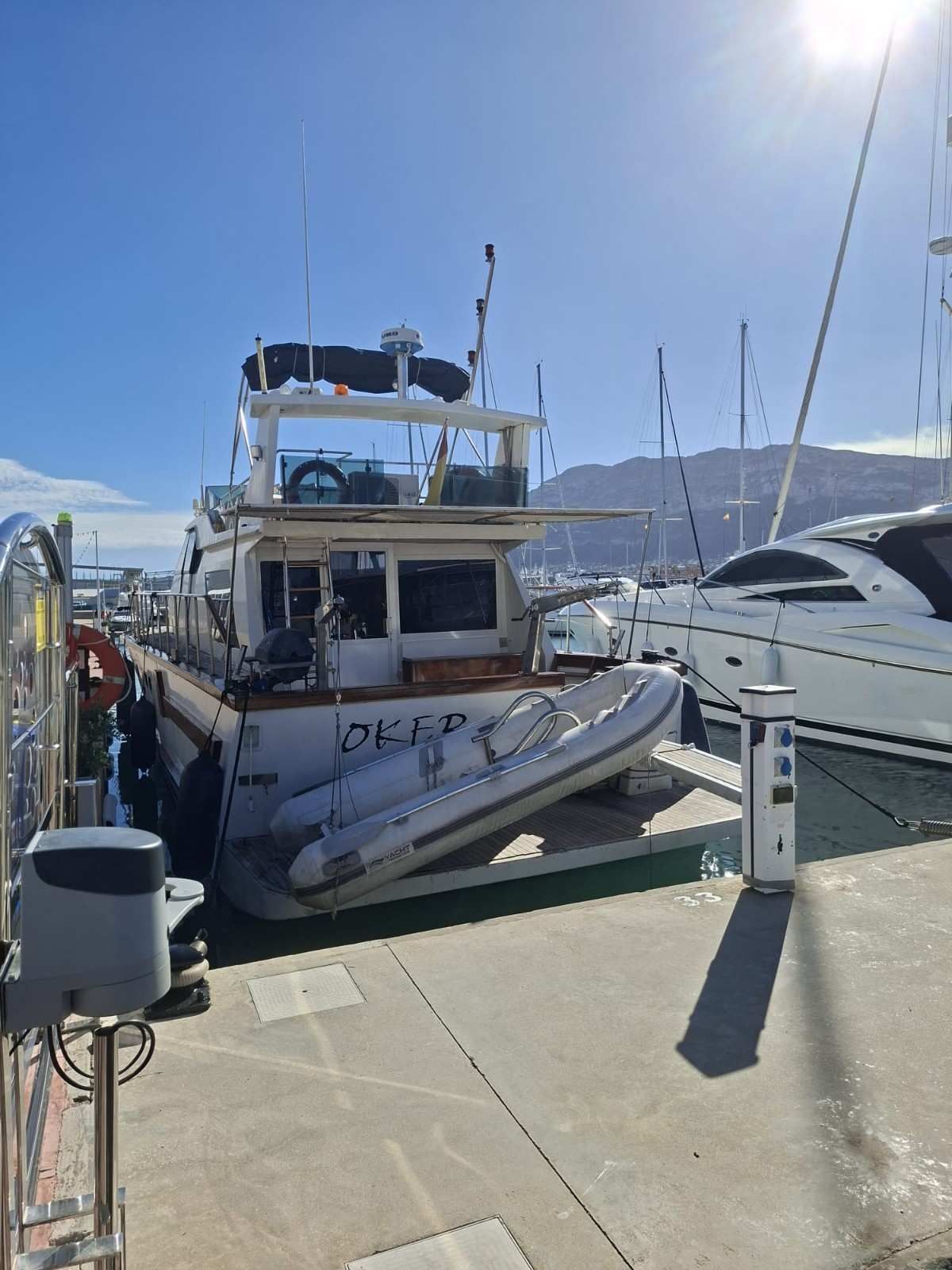 A white motor yacht with a small inflatable dinghy at the stern, docked at a marina under a bright, sunny sky.