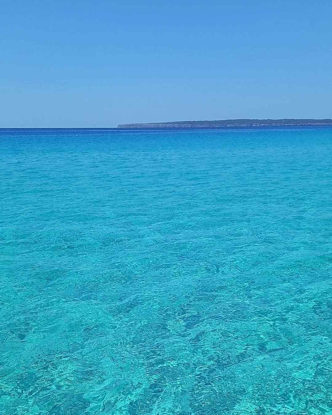 A vast expanse of crystal clear turquoise ocean water meeting a distant shoreline under a bright, cloudless blue sky.