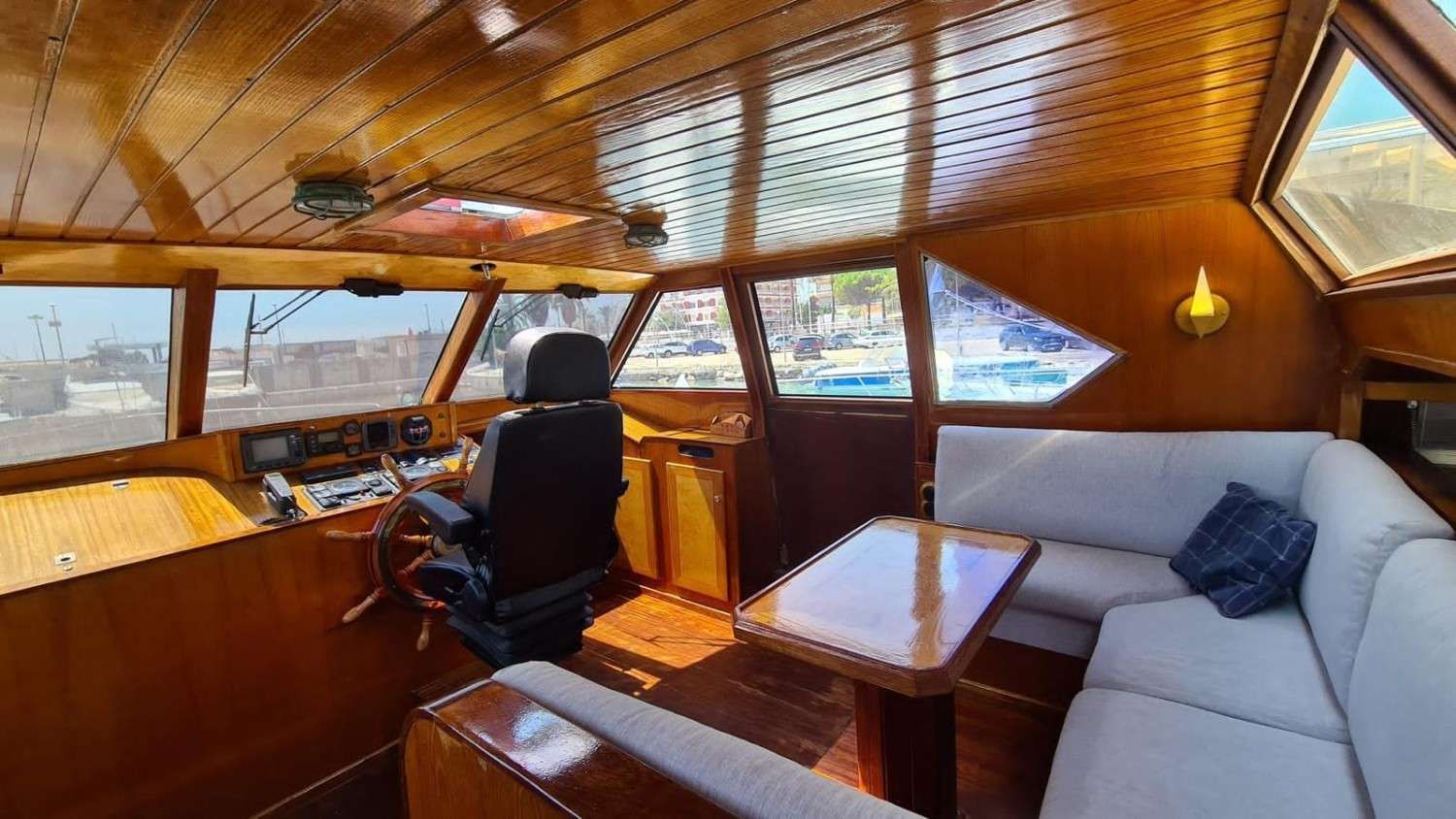 A wood-paneled yacht cabin featuring a helm station with a black captain’s chair and a lounge area with light gray seating.