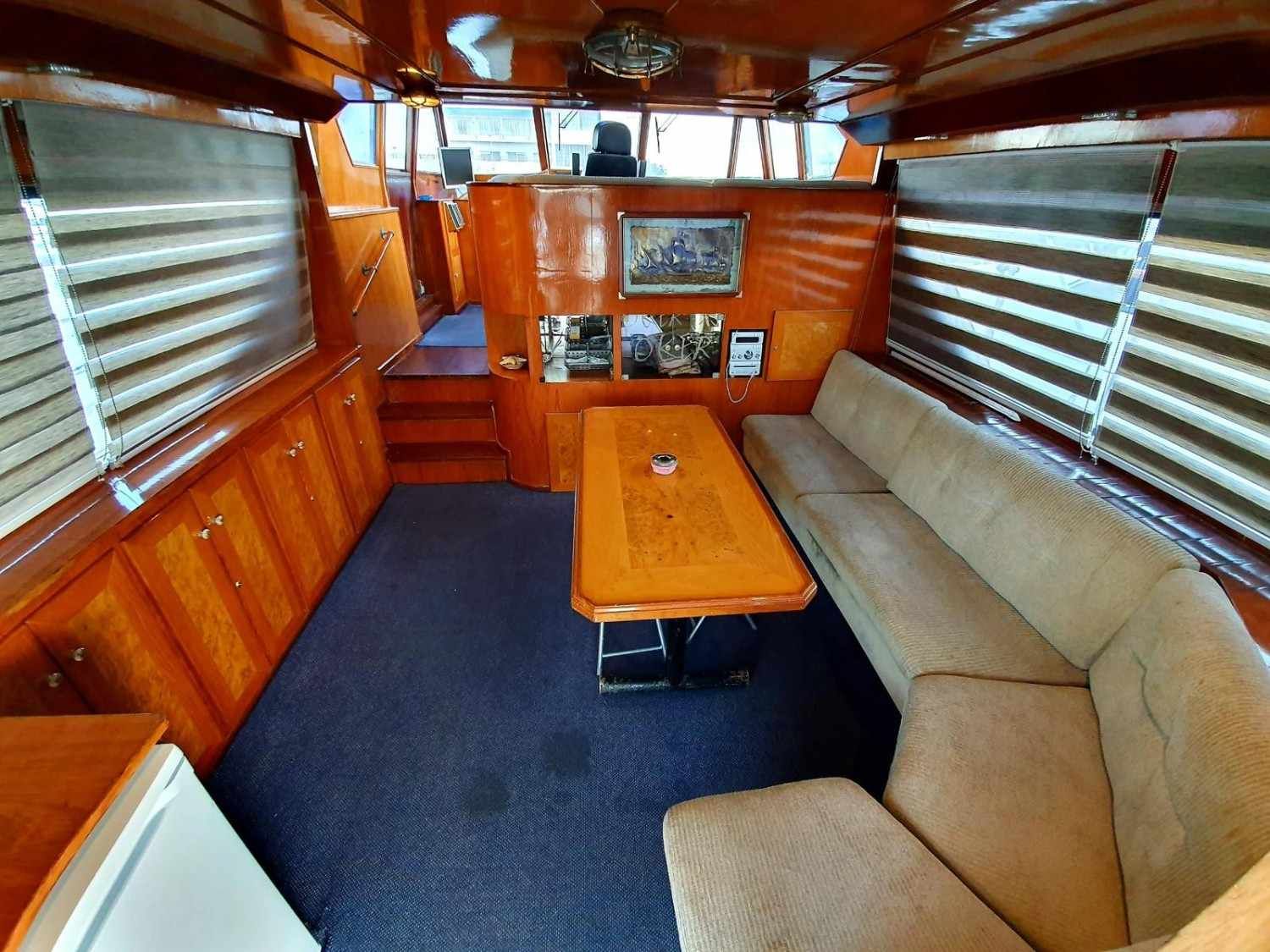 The interior of a wooden boat cabin featuring a dining table, a beige sectional sofa, dark blue carpet, and window blinds.