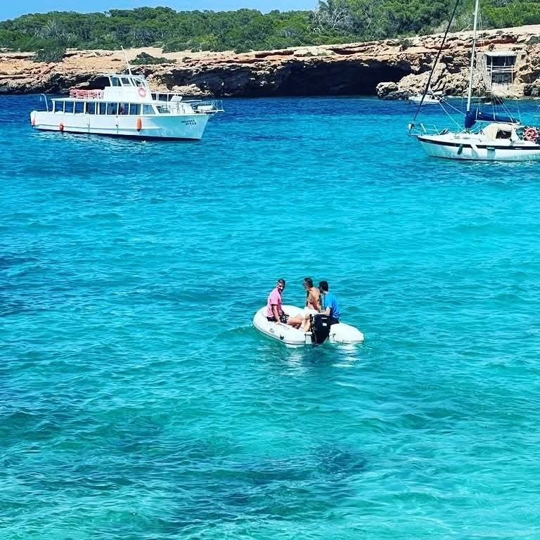 Three people in a small inflatable boat navigate turquoise waters near a rocky coastline, with two larger boats nearby.