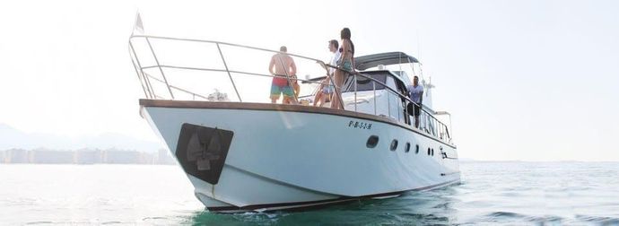 A white motor yacht carrying passengers on the deck floats on calm water under a bright sky.
