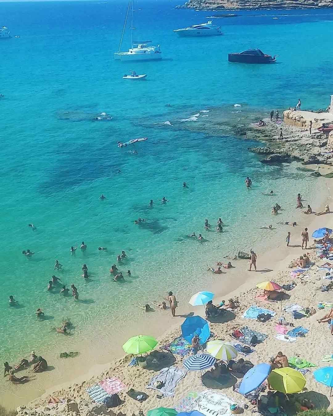 A high-angle view of a crowded, sandy beach with turquoise water, colorful umbrellas, and several boats offshore.