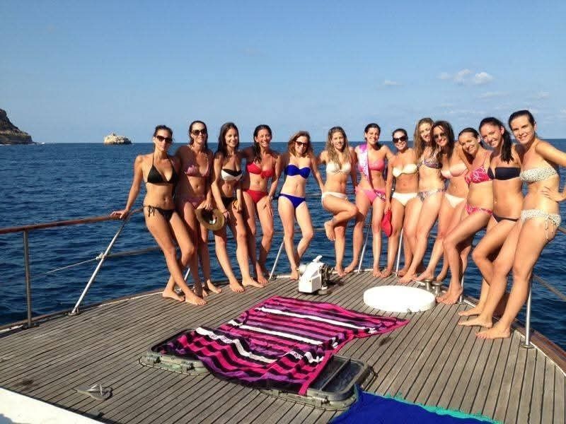 A group of women in bikinis standing on a wooden boat deck in front of the ocean on a sunny day.