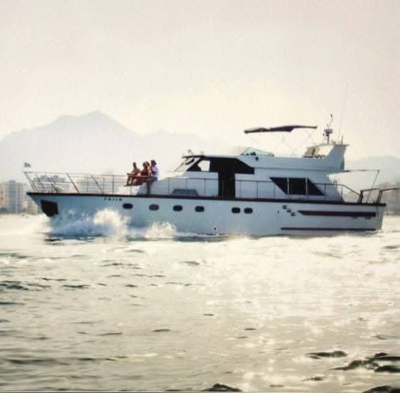 A white motor yacht cruising on the water with a group of people sitting on the deck, against a hazy mountain backdrop.