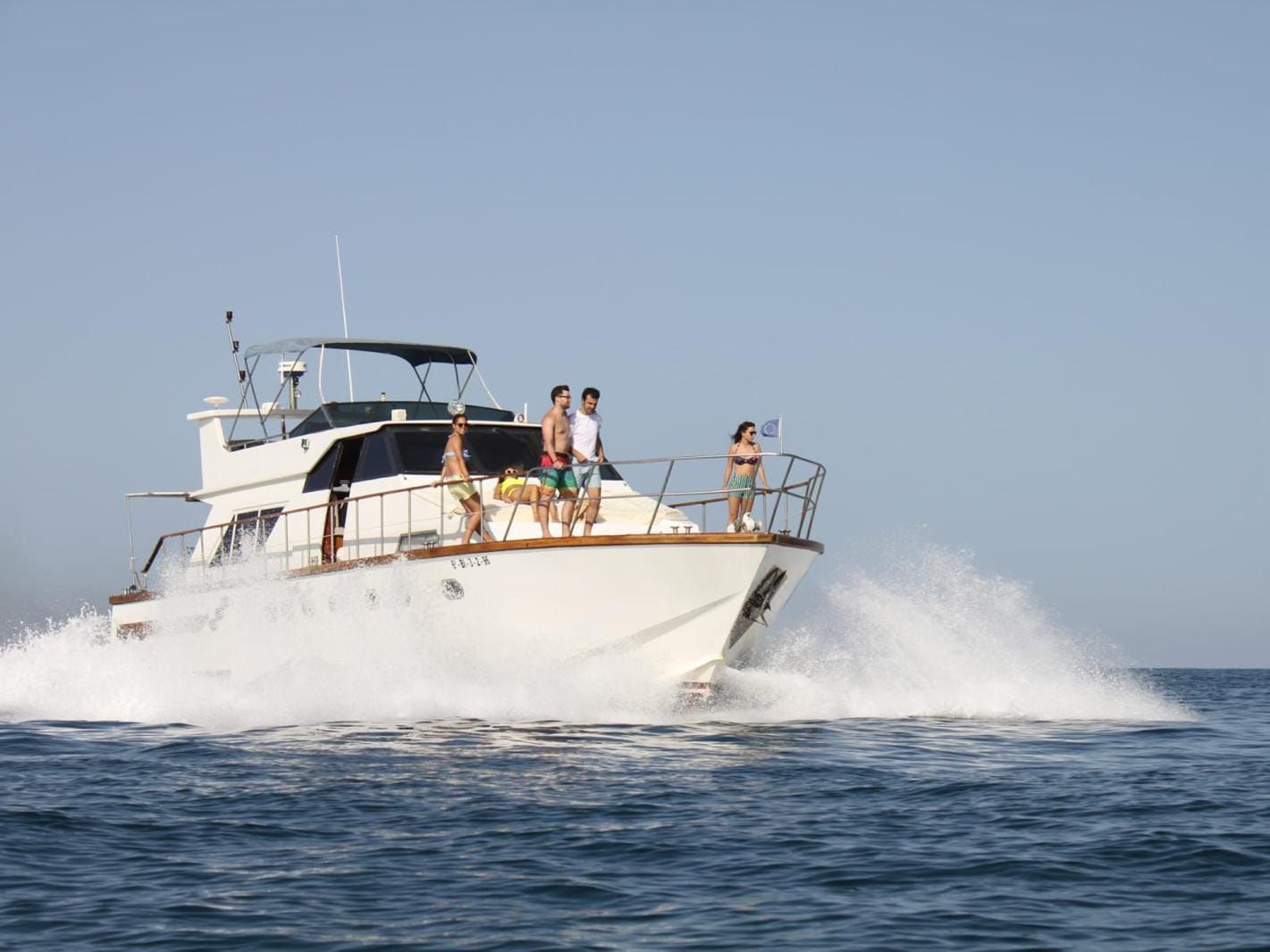 A white yacht cruises on the open ocean, creating a large white wake, with four people standing on the front deck.