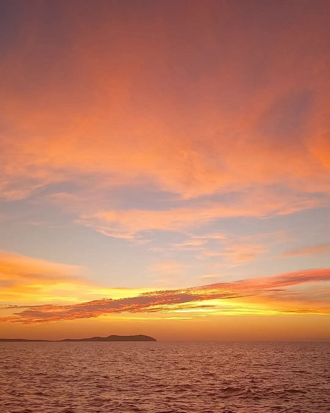 Golden sunset clouds glow over a calm ocean, with a distant, faint island silhouette on the horizon.