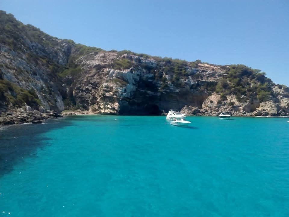Turquoise water in a coastal cove surrounded by rocky cliffs, with two white yachts anchored in the distance.