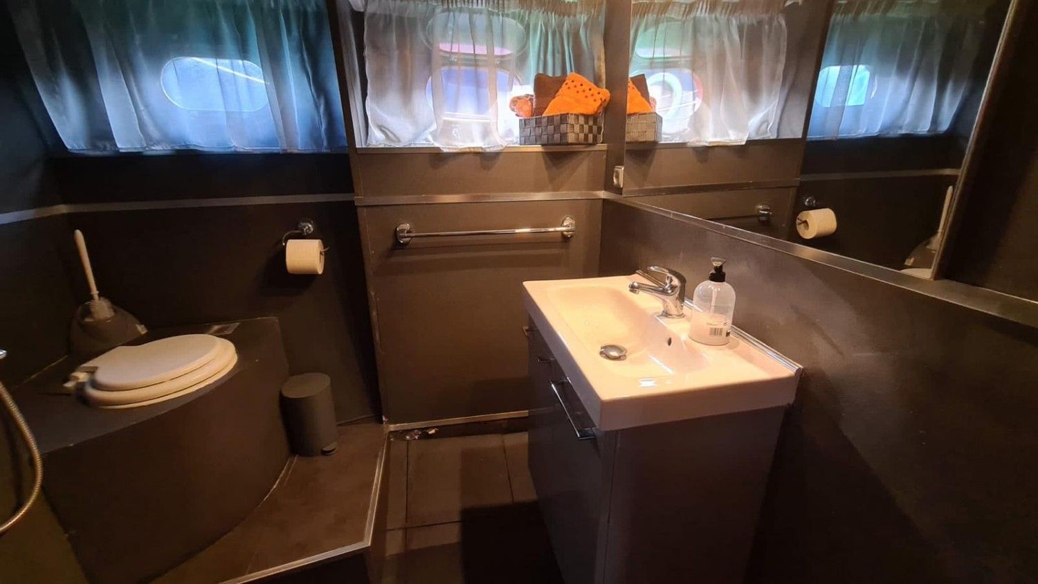 A compact bathroom with dark walls, a white pedestal sink, a toilet on a raised platform, a mirror, and window curtains.