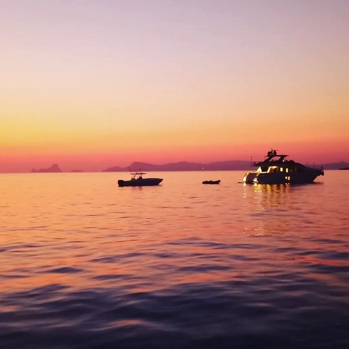 A yacht and a smaller boat silhouetted against a vibrant orange and pink sunset over calm water.