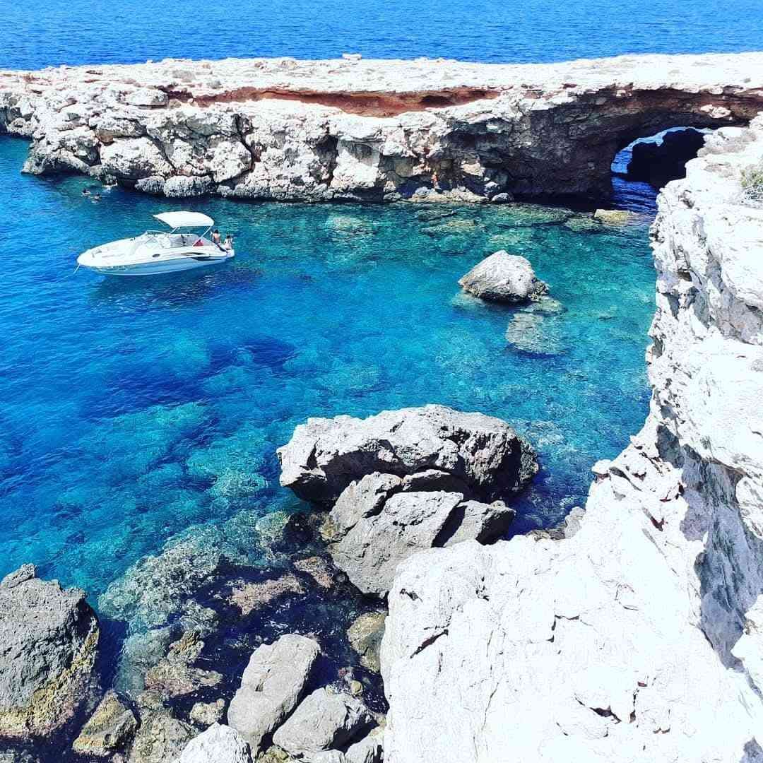 A small motorboat drifts in a bright turquoise bay surrounded by rocky cliffs and a natural stone arch.
