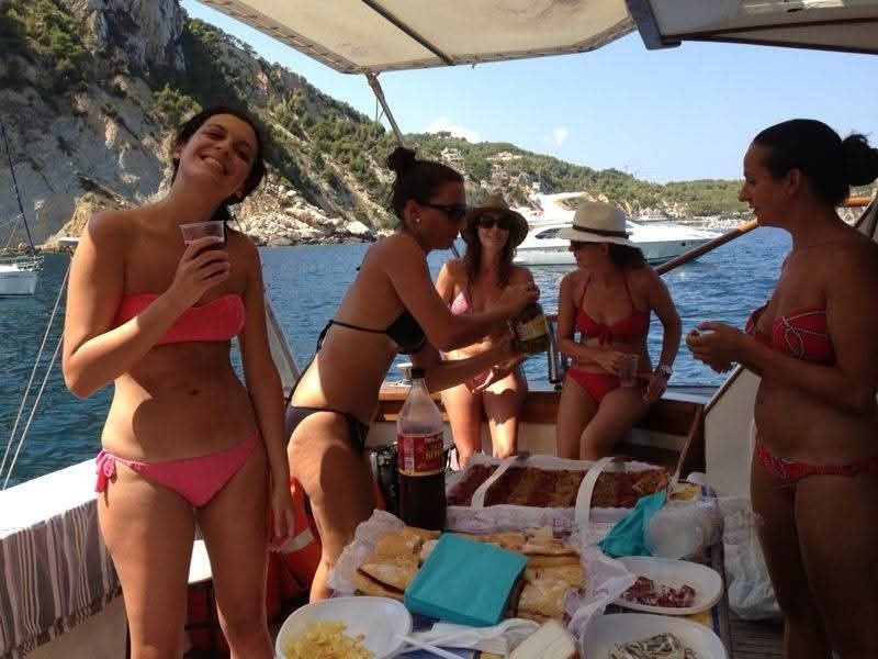 Four women in swimwear gather on a boat in a sunny, coastal setting, chatting near a table of food and drinks.