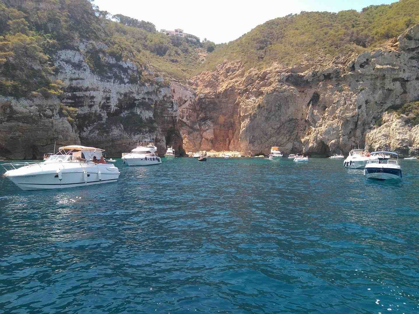Several white motorboats anchored in a calm blue bay surrounded by high, rocky cliffs under a clear sky.