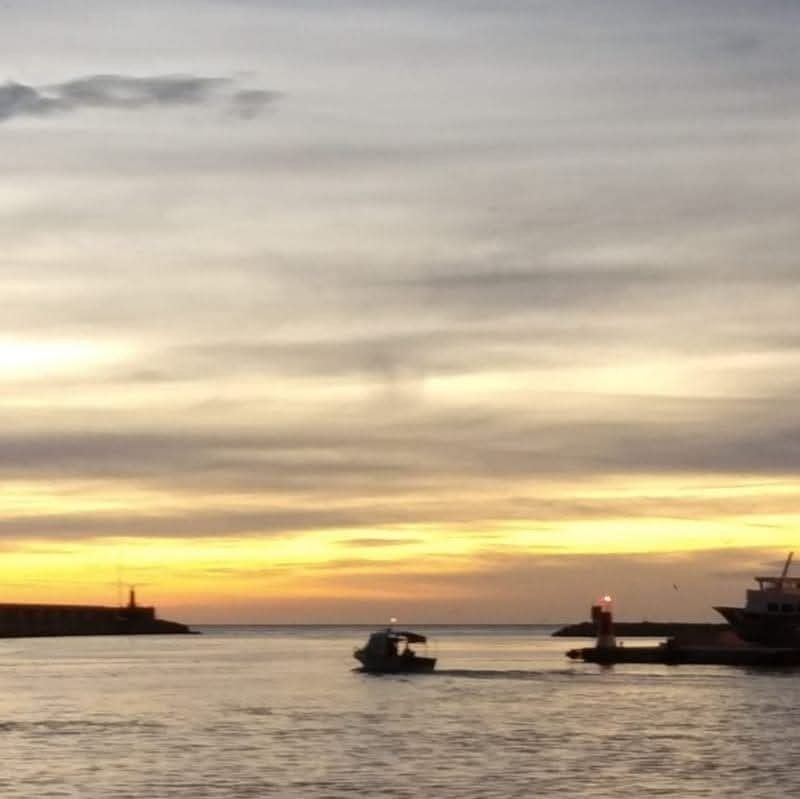 A small boat travels through a harbor entrance at sunset with soft orange, yellow, and grey skies overhead.