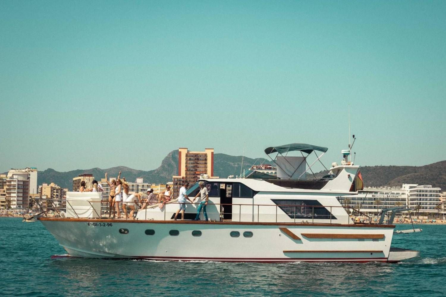 A white yacht with people on deck sailing on calm water near a coastal city with mountains in the background.