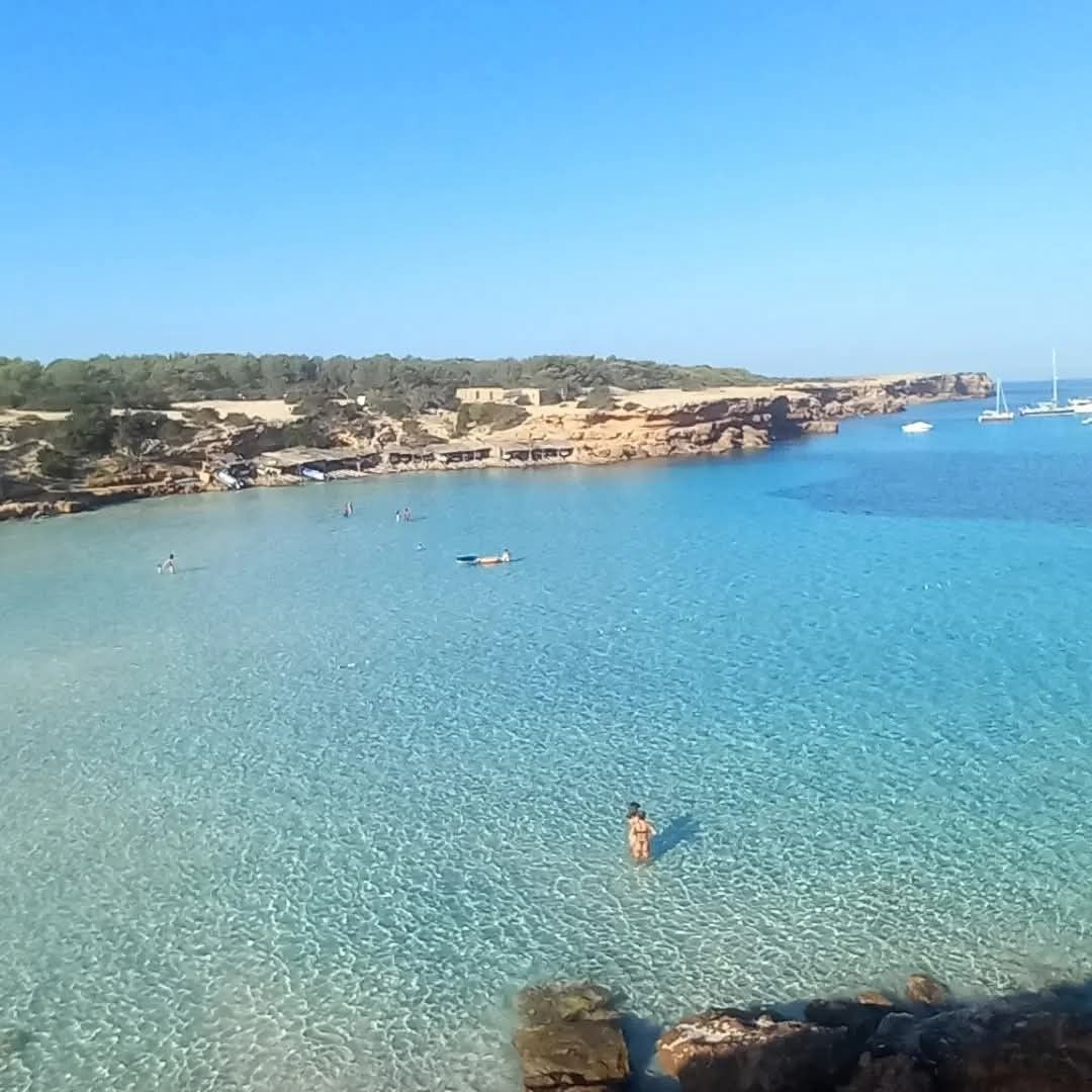 A turquoise bay with calm, clear water, a sandy beach, low rocky cliffs, and distant boats under a bright blue sky.