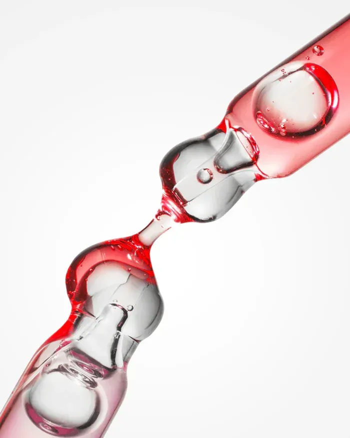 Dos goteros de vidrio que contienen y gotean líquido rojo sobre un fondo blanco.