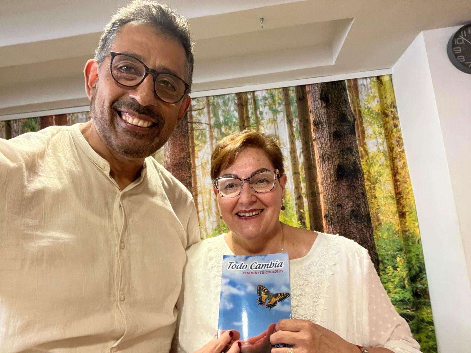 Dos personas sonriendo, sosteniendo un libro, con un bosque de fondo.