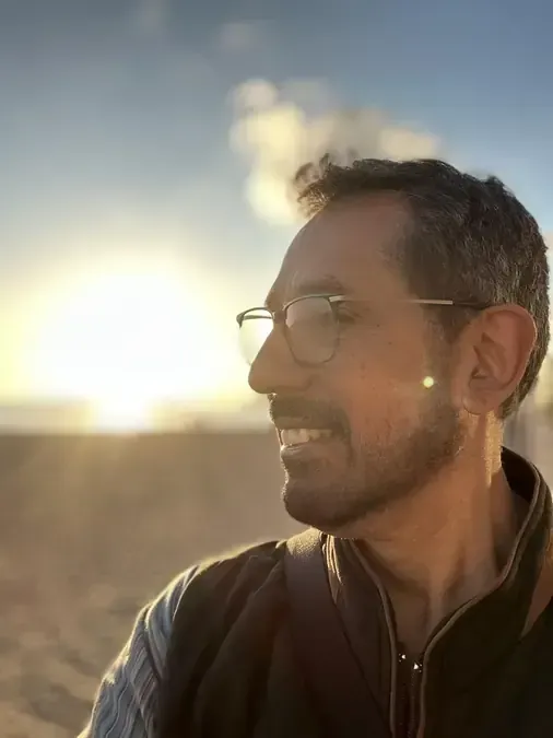 Hombre con gafas sonriendo, mirando hacia la puesta de sol en una playa.