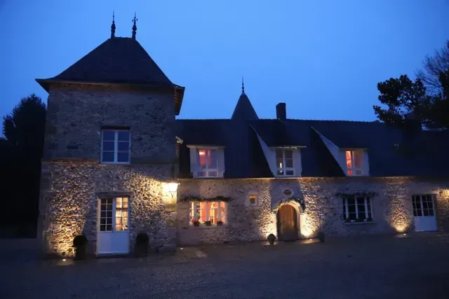 Maison de pierre chaleureusement éclairée au crépuscule, avec un ciel bleu et des fenêtres scintillantes.