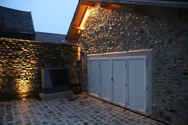 Cour en pierre éclairée par des lumières extérieures chaleureuses, avec des portes de garage blanches et un sol carrelé au crépuscule
