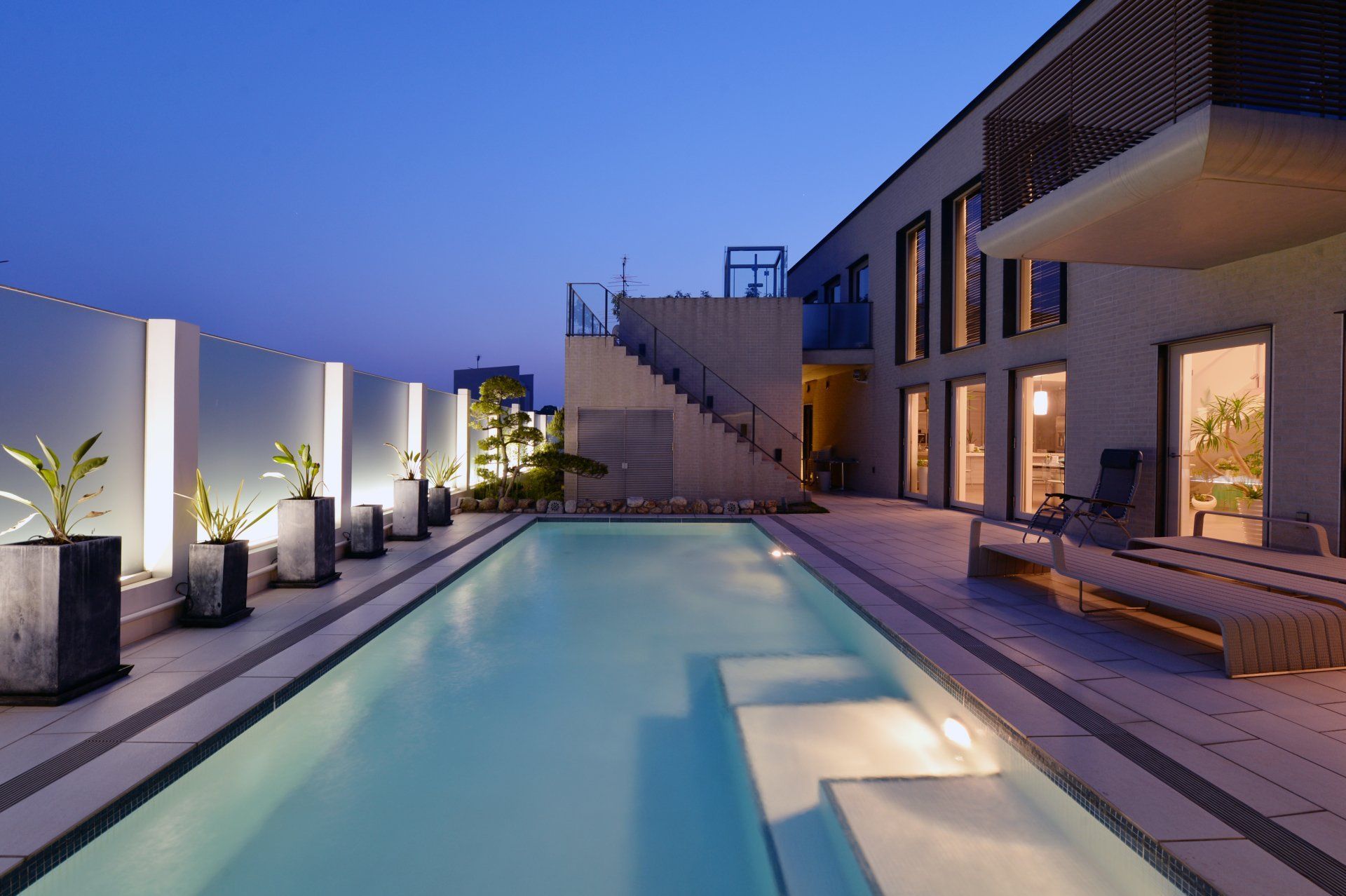 Terrasse luxueuse et moderne avec piscine illuminée au crépuscule, chaises longues et élégants murs extérieurs blancs.