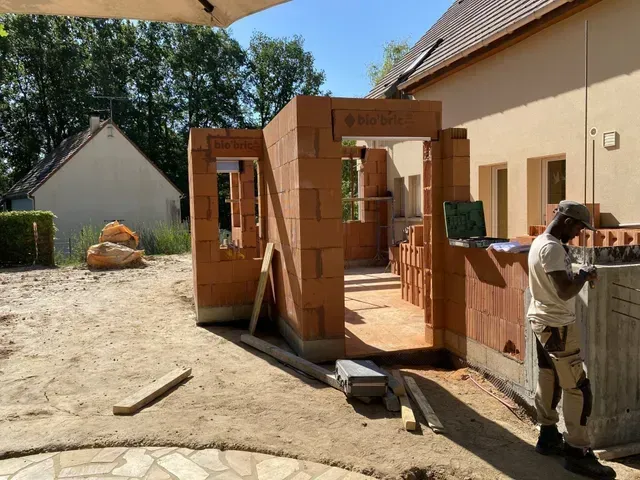 Structure en bois partiellement construite à l'extérieur, avec un ouvrier se tenant à côté, près d'une maison.