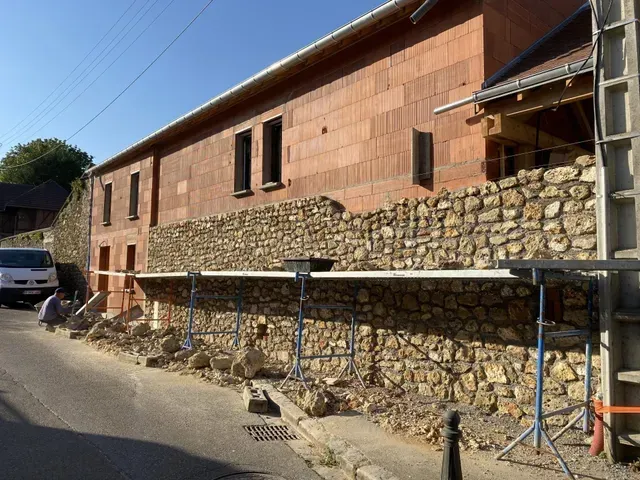 Bâtiment rural avec un mur inférieur en pierre et un étage supérieur en briques inachevé, situé le long d'une route étroite, avec des échafaudages devant.