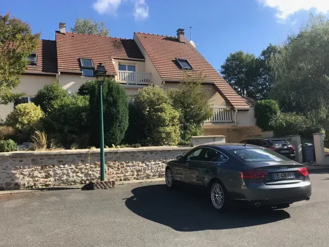 Rue résidentielle avec une voiture grise garée devant une maison au toit de tuiles rouges et entourée d'arbres.