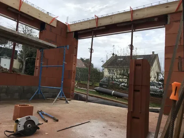 Intérieur d'un chantier de construction avec une structure métallique, des outils au sol et une vue sur les maisons à l'extérieur.
