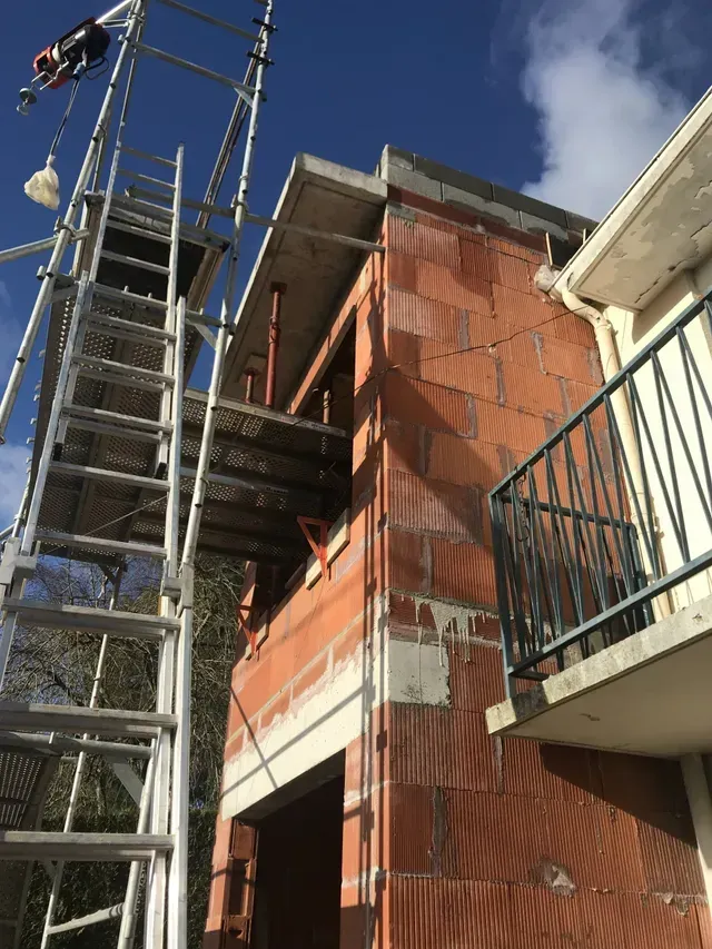 Échafaudages à côté d'un bâtiment en briques rouges en construction, avec un balcon et un ciel bleu.