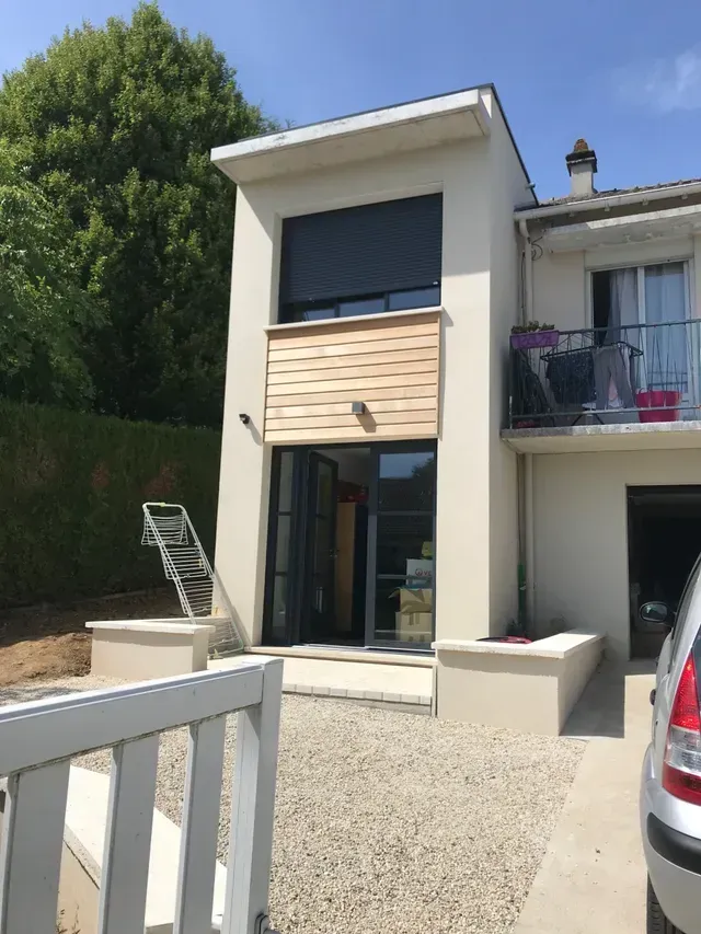 Maison moderne à deux étages avec balcon, allée gravillonnée et porte vitrée ouverte baignée de soleil.