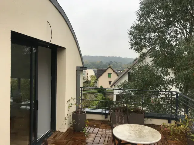 Balcon avec table ronde, chaises, plante en pot et vue sur les maisons et les arbres d'une colline brumeuse