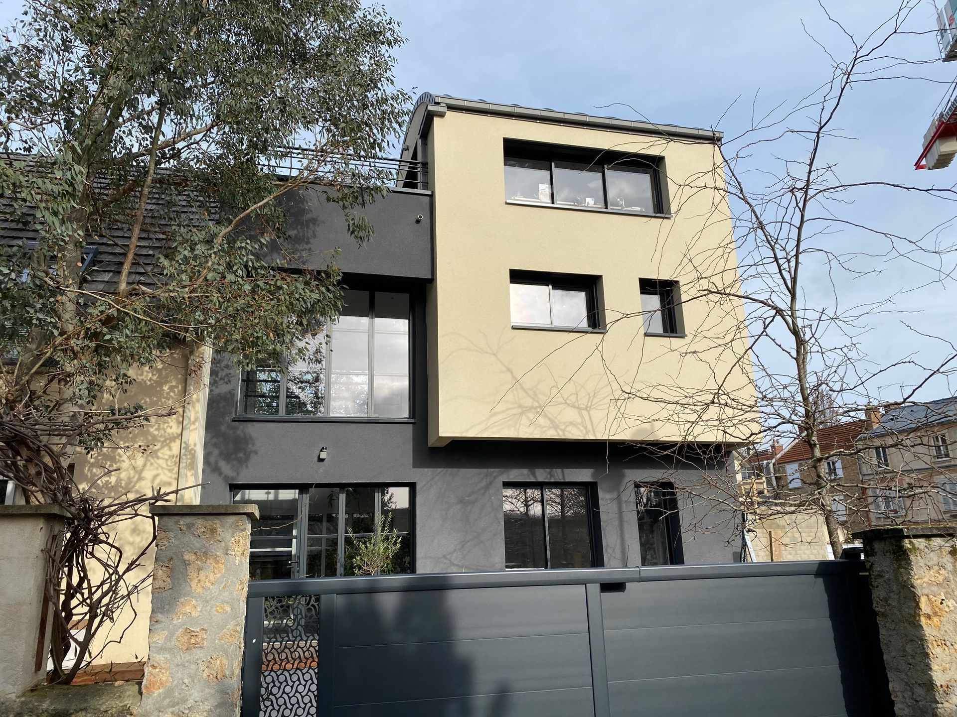 Maison moderne à deux étages avec extérieur gris foncé et beige, grandes fenêtres et portail d'entrée.