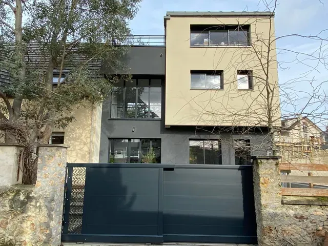Maison moderne à deux étages avec une façade beige et grise, derrière un portail bleu foncé et des arbres.