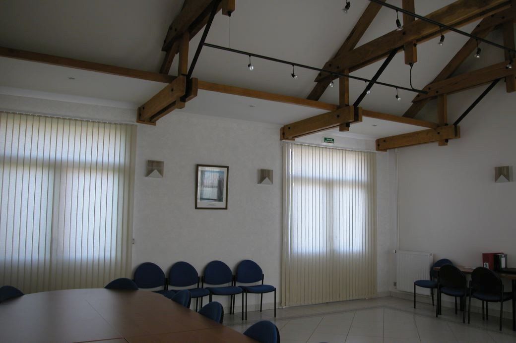 Salle de réunion lumineuse avec des rangées de chaises bleues, de hautes fenêtres à rideaux et des poutres apparentes au plafond.