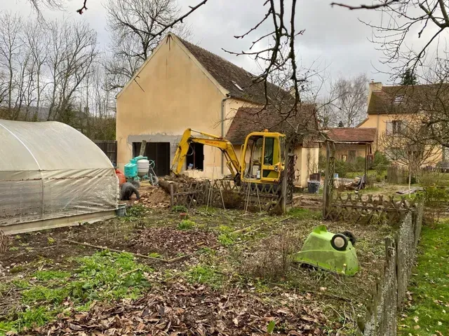 Une personne et une petite pelleteuse travaillent à côté d'une serre dans un jardin boueux.